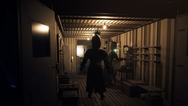 In-game: inside the crew's van under bare bulbs, one crewmate walks in while another, lens glowing verdigris, reaches across the ledger desk beside the kit rack.